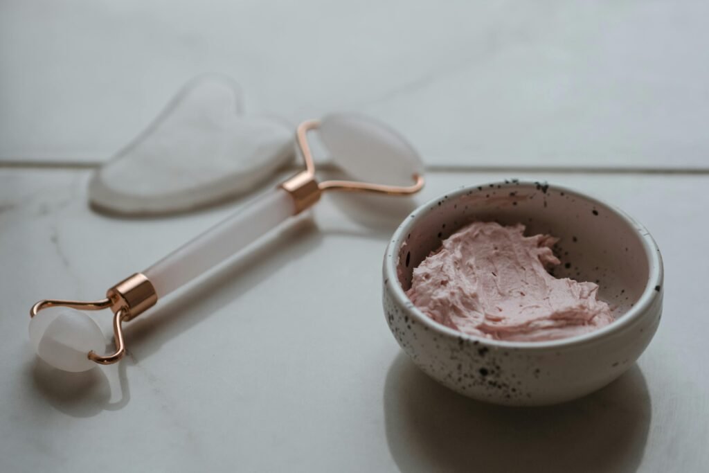Close-up of a skincare setup featuring a gua sha tool, jade roller, and pink cream in a bowl.